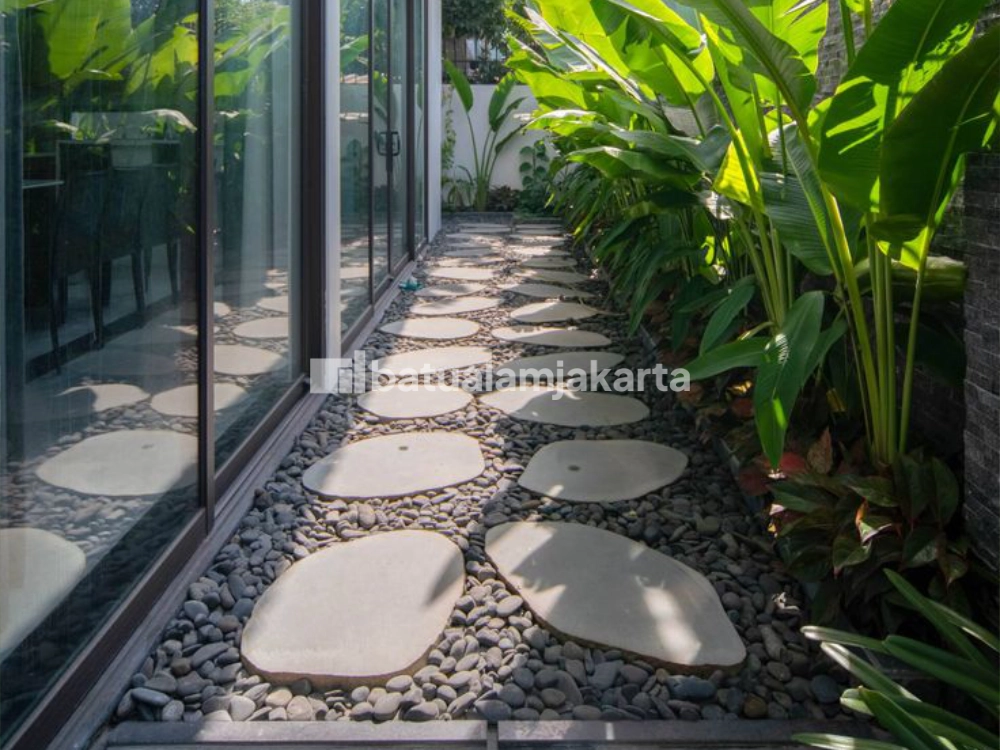 Stepping batu kali alami pada jalur taman tropis di samping bangunan.